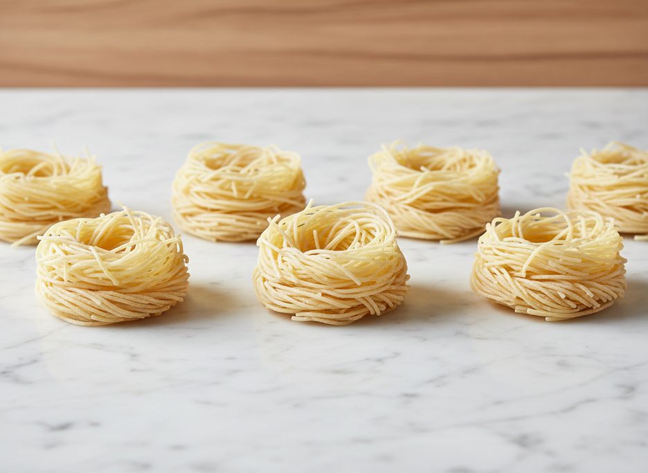 Seven small, dry, pale yellow capellini pasta nests rest on a white marble surface.