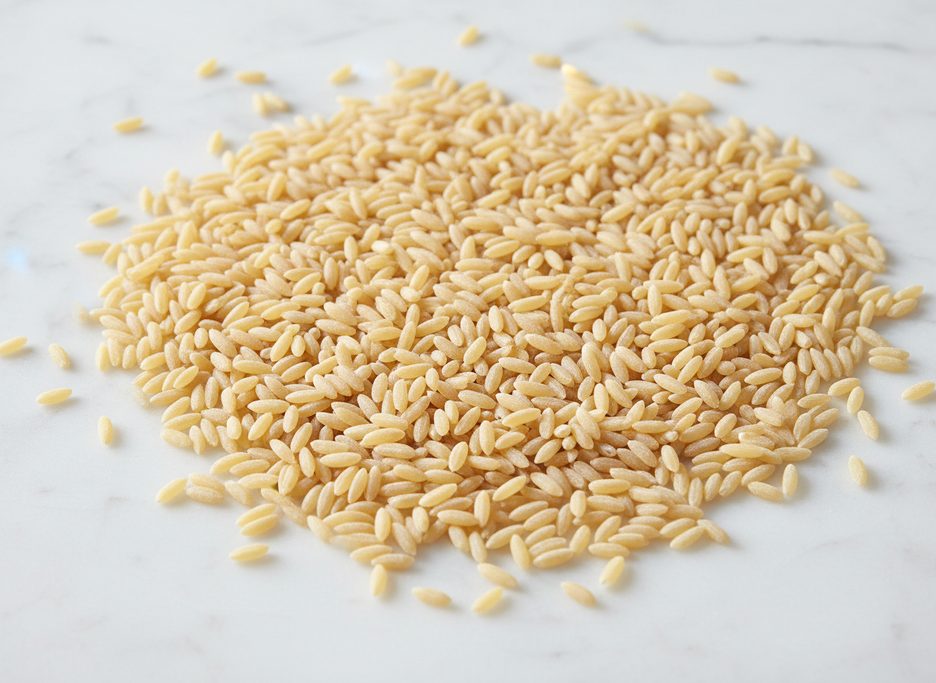 Dry, uncooked orzo pasta, small and rice-shaped, piled on a white marble surface.