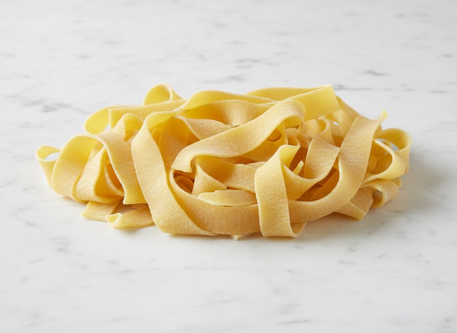 Pile of fresh, uncooked tagliatelle pasta ribbons, lightly dusted with flour, on a white marble countertop.