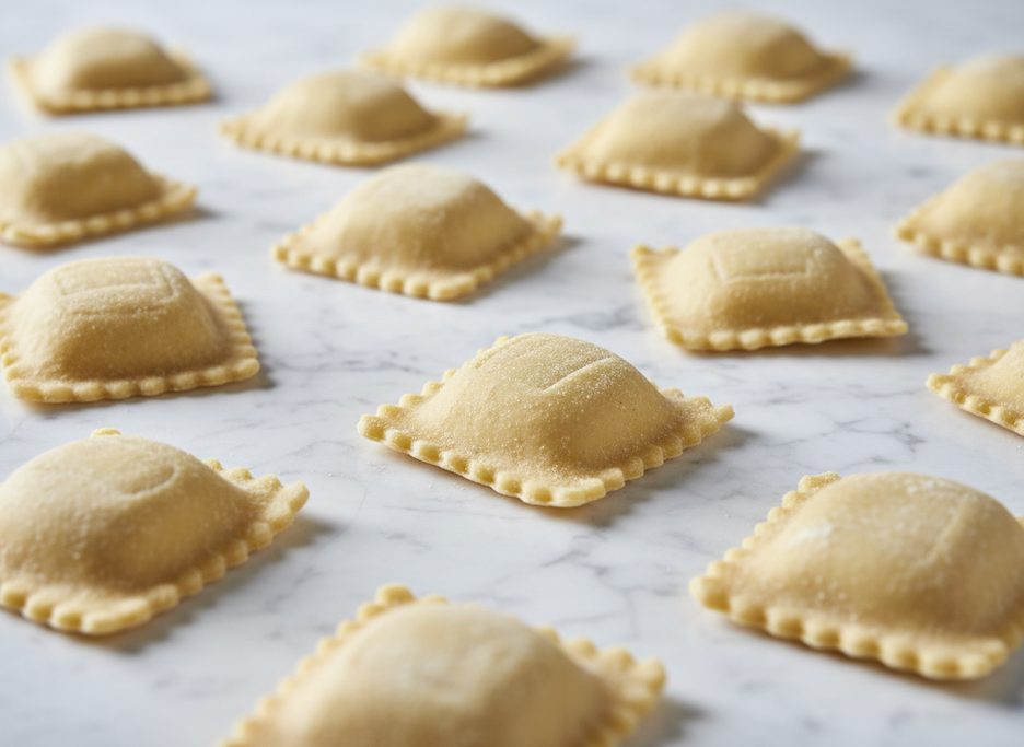 Raw square ravioli with crimped edges and a slight dome, dusted with semolina flour, on a white marble background.